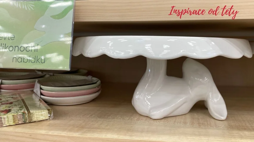Porcelánový podnos na dort ve tvaru zajíčka, který leží na zádech a nohama podpírá talíř. Podnos je umístěn na polici v Pepcu vedle velikonočních talířků.